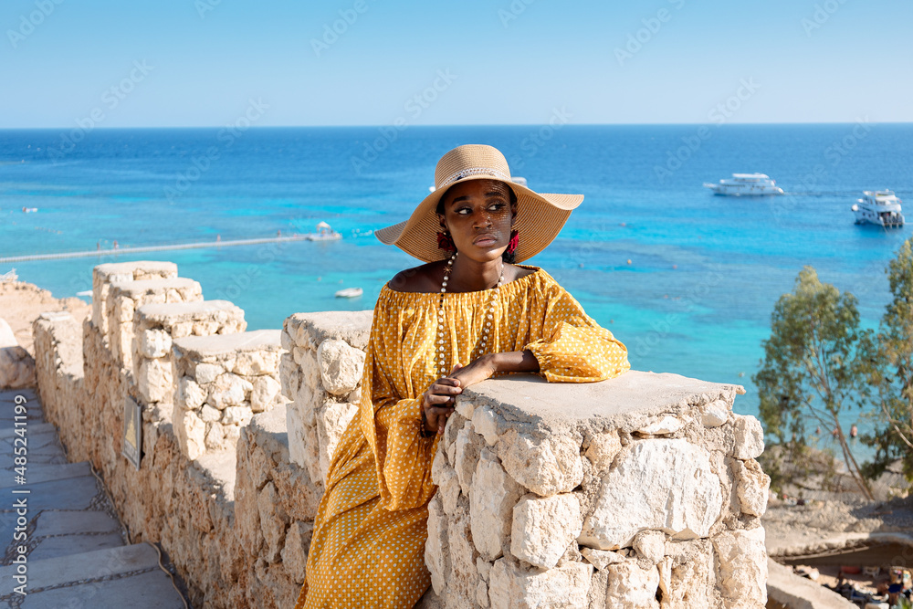 Happy African American woman in yellow dress and sun hat enjoys view of coast of Red Sea on natural background. Panoramic views of blue sea with yachts and coastline, Sharm El Sheikh, Egypt