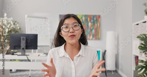A pretty smiling Korean Asian woman with glasses sits in an office dressed in a smart white shirt, a businesswoman holds a video call, conferences, asks her boss questions over the internet