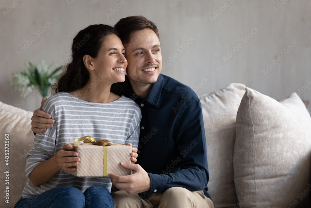 Happy young couple celebrating special event, sitting on couch, dreaming looking in distance, loving boyfriend hugging girlfriend, congratulating with anniversary or birthday, holding gift box Happy young couple celebrating special event, sitting on couch, dreaming looking in distance, loving boyfriend hugging girlfriend, congratulating with anniversary or birthday, holding gift box