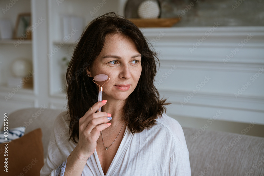 Brunette smiling woman 35 years old plus clean fresh face and hands with long hair makes facial massage with gouache roller in a bathrobe while sitting at home on the couch. Facial skin care concept Brunette smiling woman 35 years old plus clean fresh face and hands with long hair makes facial massage with gouache roller in a bathrobe while sitting at home on the couch. Facial skin care concept