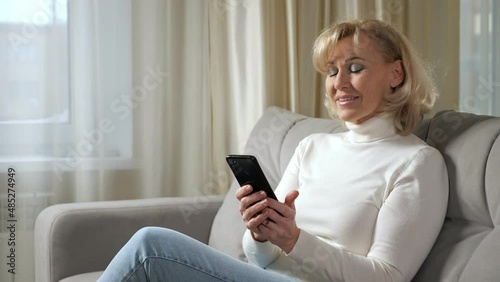 Middle aged woman with blonde bangs in poloneck smiles and scrolls through social media via smartphone on comfortable sofa in living room closeup. photo