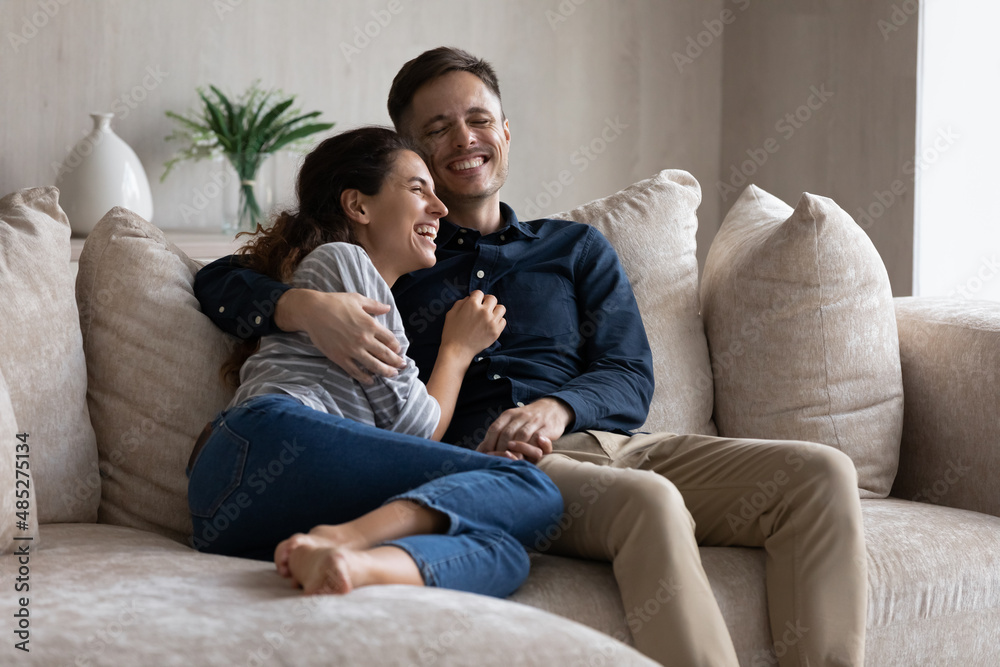 Excited couple hugging, relaxing sitting on couch having fun together, happy beautiful girlfriend and boyfriend spending leisure time lazy weekend at home, talking and laughing at funny joke Excited couple hugging, relaxing sitting on couch having fun together, happy beautiful girlfriend and boyfriend spending leisure time lazy weekend at home, talking and laughing at funny joke