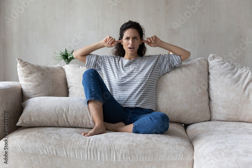 Annoyed unhappy Hispanic woman plugging ears with fingers, sitting on couch at home, noisy disturbing neighbors concept, irritated young female ignoring avoiding loud sound of repairmen