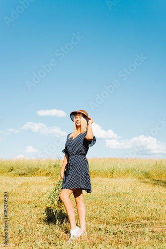 Pretty pretty young woman posing in summer dress, straw hat and with bouquet of wild flowers on sunny day in meadow outdoors. Cute smiling caucasian girl standing on field and looking away, lifestyle