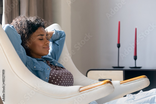 Billede på lærred Smiling peaceful young African American woman relaxing in comfortable armchair with folded arms behind head, breathing fresh air, sleeping napping enjoying weekend leisure time, meditating at home