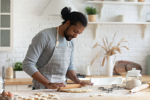Happy handsome young 30s African American man in apron rolling out dough for homemade pastry, enjoying preparing biscuit cookies in modern light kitchen, cooking hobby activity pastime concept. photo
