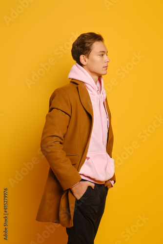 Handsome stylish man in brown trenchcoat and pink sweatshirt with hood standing in profile over yellow background photo