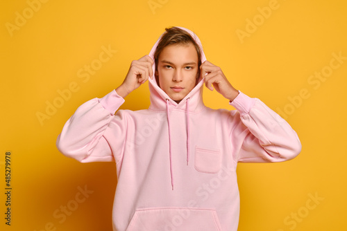 Thoughtful young man in pink sweatshot puts on hood over yellow background photo