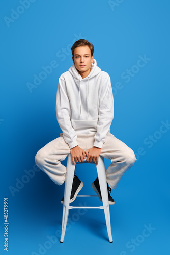 Handsome man wearing white sweatshirt and sweatpants sitting on white chair over blue background photo