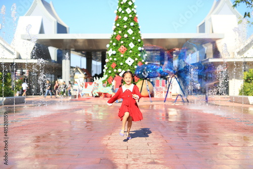 Happy little Asian girl in red dress running around the big christmas tree decorative for the happy new year and merry christmas festival.
