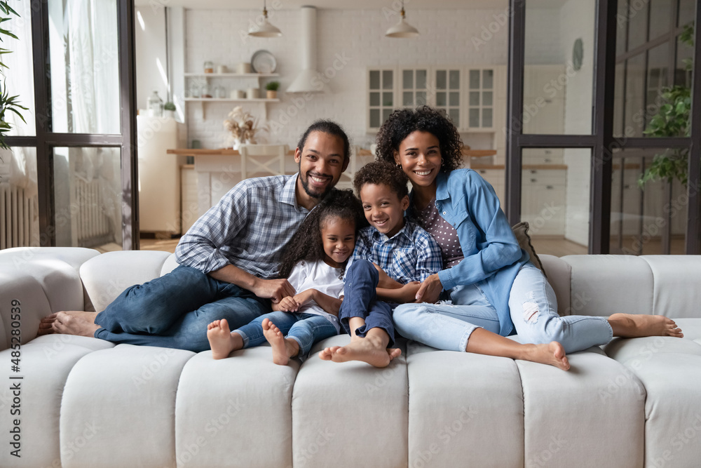 Full length portrait of smiling friendly African American family with cute little kids siblings relaxing on huge cozy couch in modern living room, enjoying carefree weekend time together at home. Full length portrait of smiling friendly African American family with cute little kids siblings relaxing on huge cozy couch in modern living room, enjoying carefree weekend time together at home.