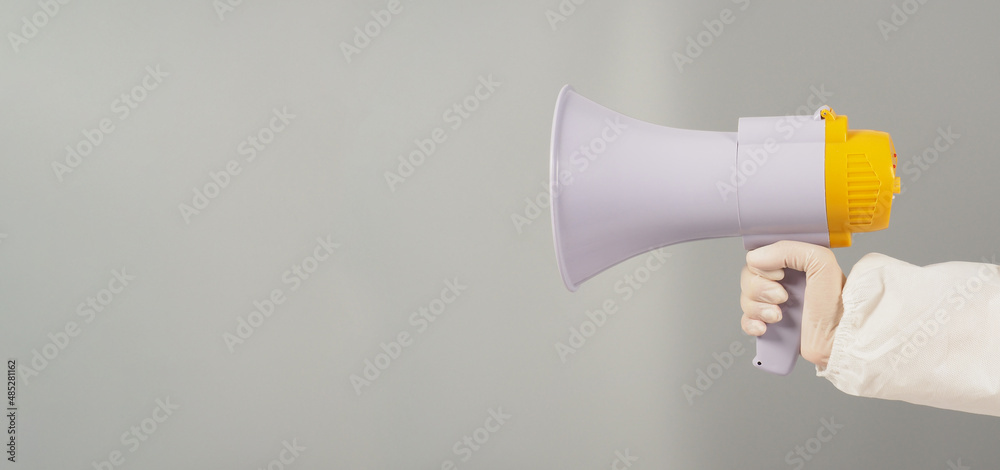 Hand is holding megaphone on grey background. Hand wears a PPE suit and a white medical glove. Hand is holding megaphone on grey background. Hand wears a PPE suit and a white medical glove.
