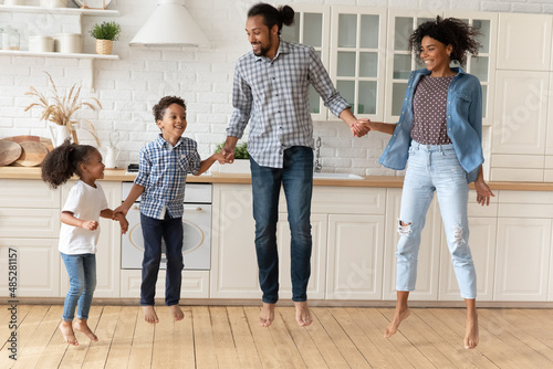 Full length happy millennial African American couple parents jumping barefoot, having fun dancing to energetic disco music together with cheerful small kids siblings in modern renovated kitchen.
