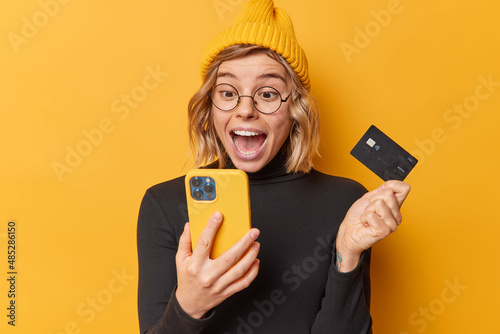 Surpised happy woman wears hat and casual black turtleneck looks at smartphone app screen holds credit card uses bank account for paying online isolated over yellow background gets cashback.