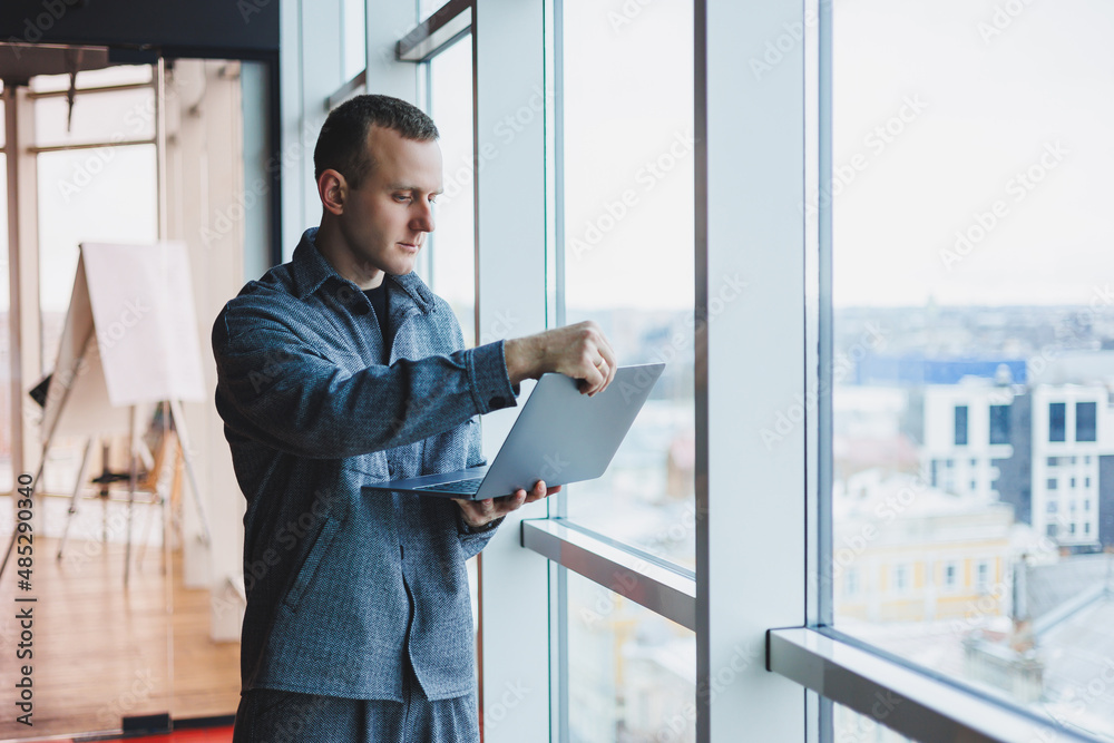 Portrait of a smart intelligent male manager, he is holding a laptop for research in the office, a business man in elegant clothes playing on the touchpad. Standing in the office by a large window Portrait of a smart intelligent male manager, he is holding a laptop for research in the office, a business man in elegant clothes playing on the touchpad. Standing in the office by a large window