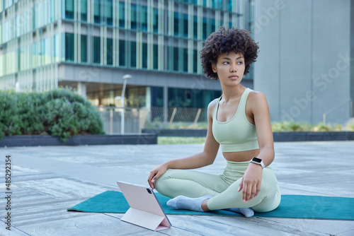 Fit healthy curly woman dressed in sportswear watches meditation video on tablet sits crossed legs on fitness mat follows online yoga lesson poses in urban setting concentrated thoughtfully away