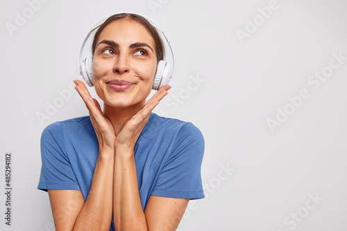 Dreamy brunette young woman has thoughtful expression keeps hands on stereo headphones wears casual blue t shirt isolated over white background blank copy space for your promotional content. photo