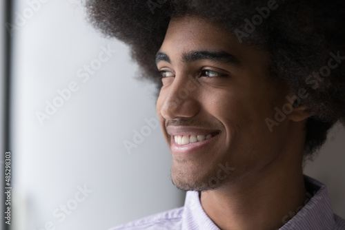 Cheerful handsome young Black guy with retro Afro hairdo looking away, dreaming with toothy smile, enjoying good happy thoughts, thinking over future vision. Dreamy African man close up portrait