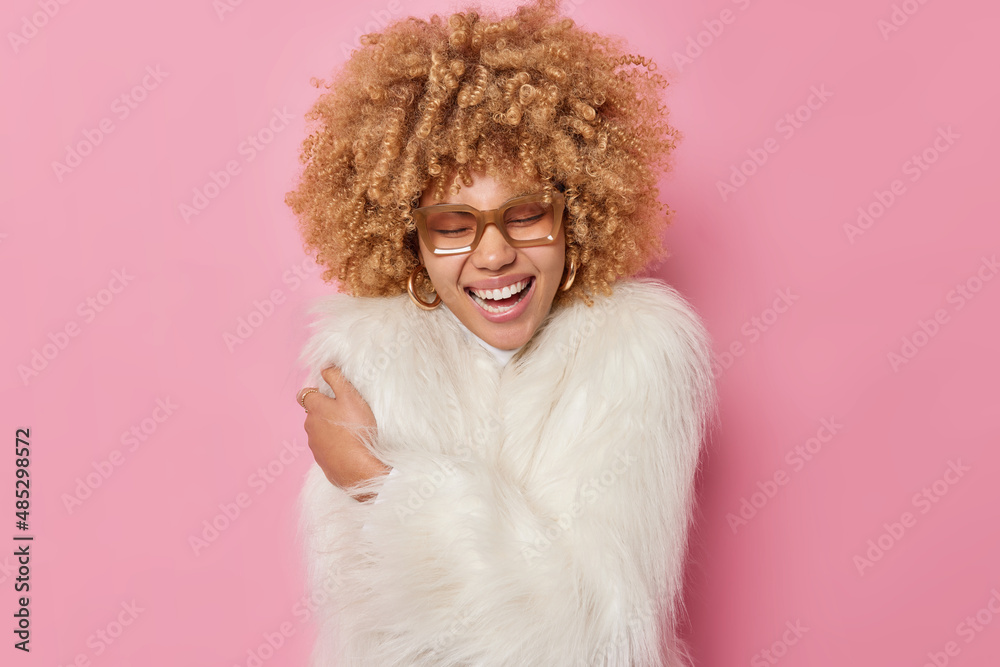 Cheerful fashionable woman with curly bushy hair feels comfortable in new winter coat embraces own body wears spectacles laughs happily isolated over pink background. People and style concept Cheerful fashionable woman with curly bushy hair feels comfortable in new winter coat embraces own body wears spectacles laughs happily isolated over pink background. People and style concept