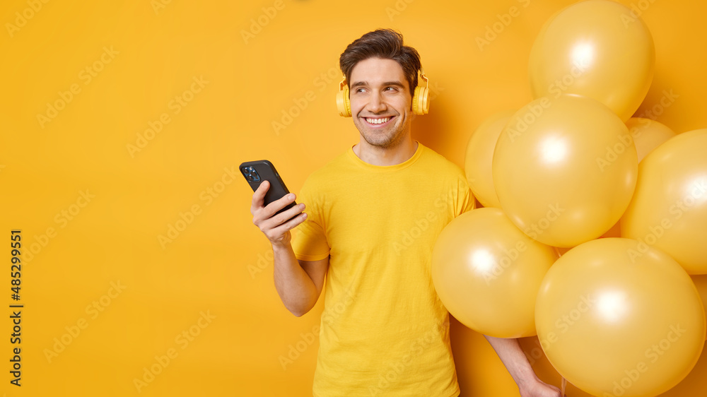 Happy dreamy man looks away with cheerful expression listens smartphone andstereo headphones for listening music holds bunch of balloons isolated over yellow background copy space for your promotion Happy dreamy man looks away with cheerful expression listens smartphone andstereo headphones for listening music holds bunch of balloons isolated over yellow background copy space for your promotion