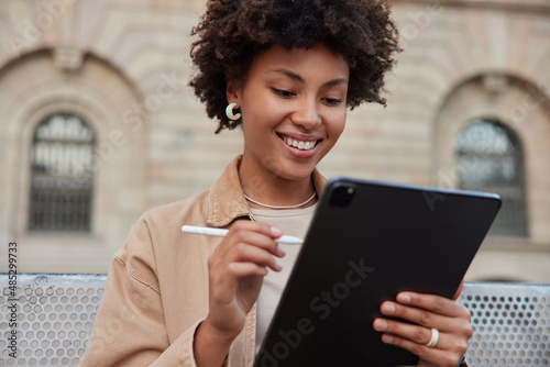 Fotografie, Tablou Positive curly woman smiles happily uses stylus and digital tablet makes creative project satisfied with results poses against ancient building being in good mood