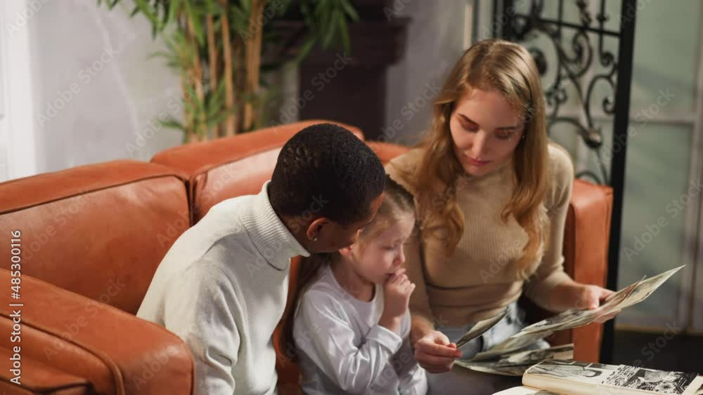 Thoughtful little girl looks at old family photos sitting with African-American father and white mother with loose hair in living room slow motion Thoughtful little girl looks at old family photos sitting with African-American father and white mother with loose hair in living room slow motion