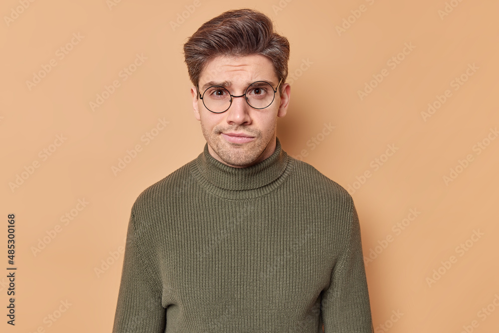 Serious worried man purses lips looks attentively at camera being dissatisfied with something weas round eyegasses for vision correction casual jumper isolated over brown background. Face expressions