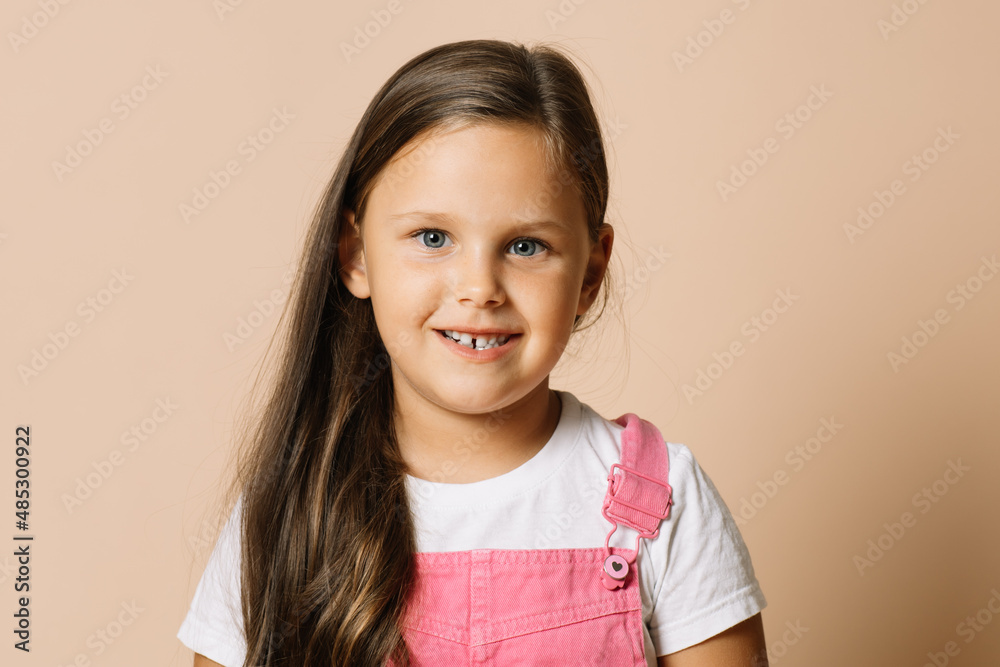 Close-up face kid with bright shining eyes and calm happy smile with teeth looking at camera wearing bright, pink jumpsuit and white t-shirt on beige background. Close-up face kid with bright shining eyes and calm happy smile with teeth looking at camera wearing bright, pink jumpsuit and white t-shirt on beige background.