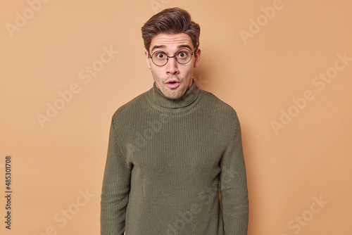 Amazed young adult man with dark hair looks wondered at camera reacts on shocking news wears round spectacles and sweater isolated over beige background. People reactions and big surprisement