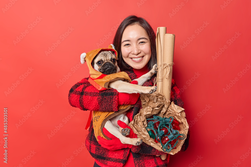 Horizontal shot of lovely cheerful dark haired Asian woman embraces pug dog dressed in winter clothes with love carries decorations for holidays has good mood isolated over vivid red background.