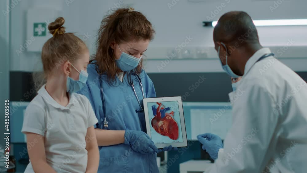 Woman nurse showing cardiology image on digital tablet, letting doctor explain cardiovascular diagnosis to child and parent. Assistant holding modern device with heart anatomy picture. Woman nurse showing cardiology image on digital tablet, letting doctor explain cardiovascular diagnosis to child and parent. Assistant holding modern device with heart anatomy picture.