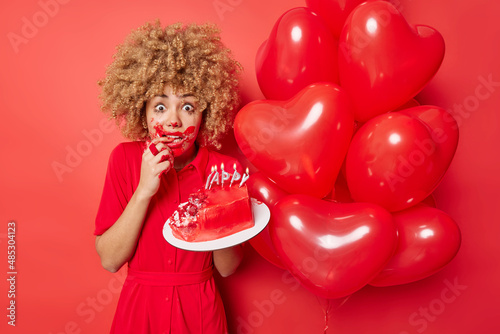 Celebration concept. Worried shocked curly woman dirty with cream holds delicious cake with burning candles bunch of inflated heart balloons prepares for lovers day. Omg Valentines is coming