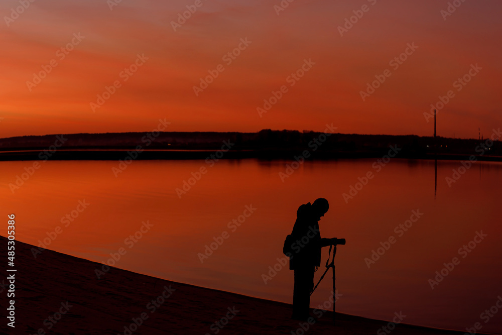 A silhouette of a photographer at sunset. A man shooting a landscape in the evening with a tripod
