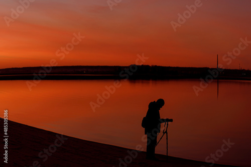 A silhouette of a photographer at sunset. A man shooting a landscape in the evening with a tripod