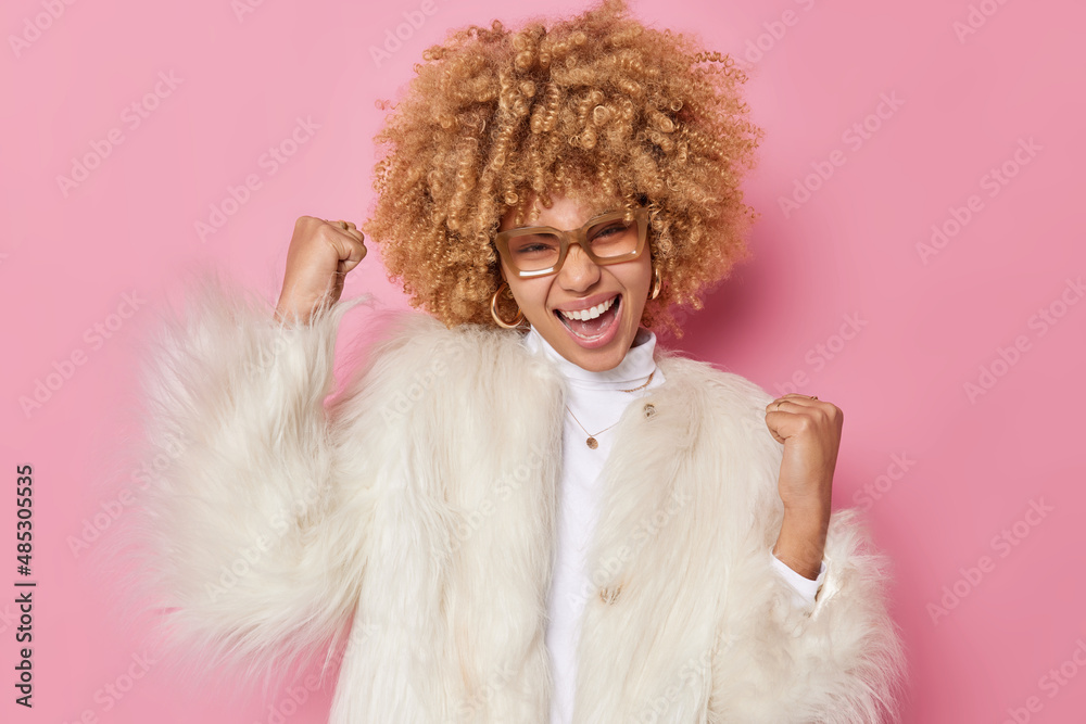 Upbeat energetic young woman with curly hair makes fist bump celebrates success exclaims from joy wears transparent spectacles and winter coat isolated over pink background. Yes I did it finally Upbeat energetic young woman with curly hair makes fist bump celebrates success exclaims from joy wears transparent spectacles and winter coat isolated over pink background. Yes I did it finally