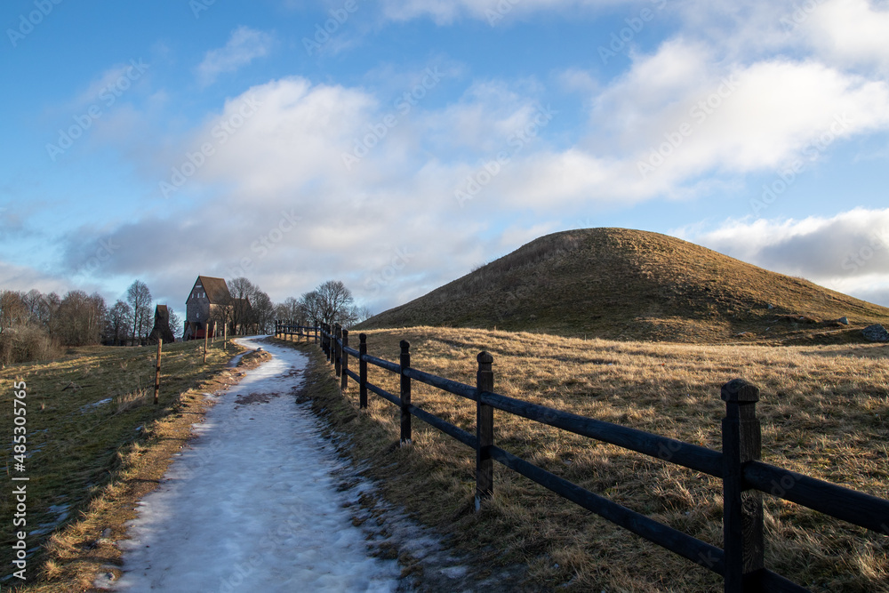 Burial mound covered with grass in culture landscape. Large oak trees and footpath with wooden fence. Ice on the ground.