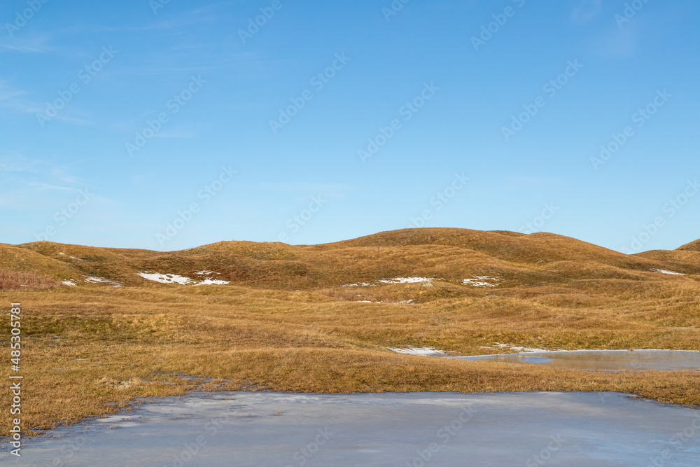 Burial mound covered with grass in rolling hill culture landscape. Patches of snow on the ground and clear sky with light clouds