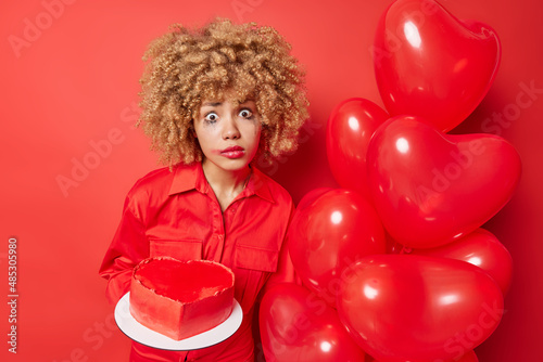 Shocked worried woman has spoiled makeup after crying feels lonely on Valentines Day holds bunch of inflated balloons and heart shaped cake sad boyfriend didnt come on date isolated on red wall