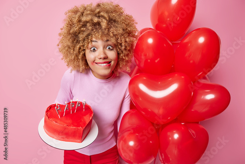 Pleased woman has hope to find her love before St Valentines Day poses with heart shaped balloons and tasty handmade cake congratulates you with coming hliday dressed casually. Celebration concept photo