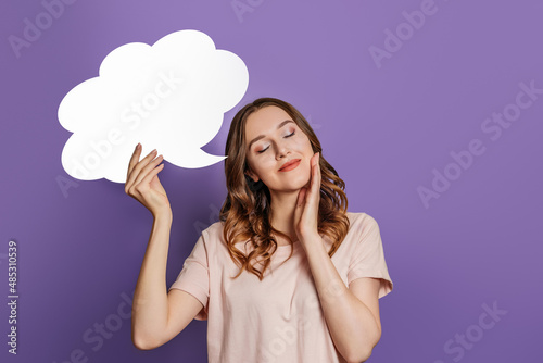 Student girl dreams with closed eyes and holding blank speech bubble isolated on purple background. Copy space