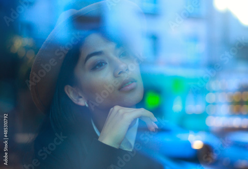 Young asian woman in classic business style sits pensively peering through window with reflections of street and lights with cup of coffee