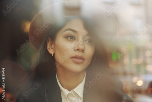 Young asian woman in classic business style sits pensively peering through window with reflections of street and lights with cup of coffee