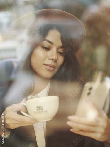 Young asian woman in classic business style sits pensively peering through window with reflections of street and lights with cup of coffee
