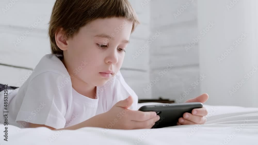 Little 5 year baby boy lays on bed and plays with smartphone. Closeup portrait. 4K video footage. Online education concept. White room and clothes. Child is holding and scrolling the telephone screen. Little 5 year baby boy lays on bed and plays with smartphone. Closeup portrait. 4K video footage. Online education concept. White room and clothes. Child is holding and scrolling the telephone screen.