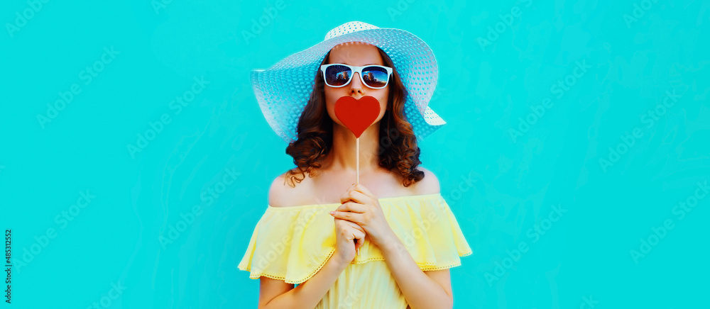 Portrait of beautiful young woman blowing her lips with lipstick with red sweet heart shaped lollipop wearing a summer straw hat on blue background