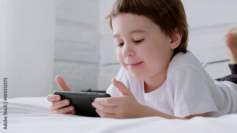 Happy 5 year boy play smartphone video game. Kid use mobile phone for gaming or learning. Smiling child in white shirt lie in home bedroom. Online education. Close-up. 4K video footage. Copy space. Happy 5 year boy play smartphone video game. Kid use mobile phone for gaming or learning. Smiling child in white shirt lie in home bedroom. Online education. Close-up. 4K video footage. Copy space.