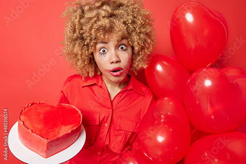 Surprised woman with fair curly hair prepares for St Valentines Day holdsheart shaped balloons and home made cake has leaked makeup poses against vivid red background. Holiday is coming soon. © wayhome.studio 