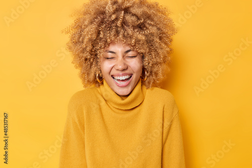Foto Photo of overjoyed European woman laughs sinecerely smiles broadly has upbeat mood keeps eyes closed wears casual warm jumper isolated over yellow background