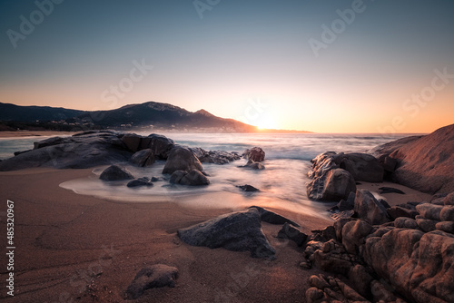 Sun setting over a rocky cove at Aregno Plage in the Balagne region of Corsica with the village of Algajola in the distance