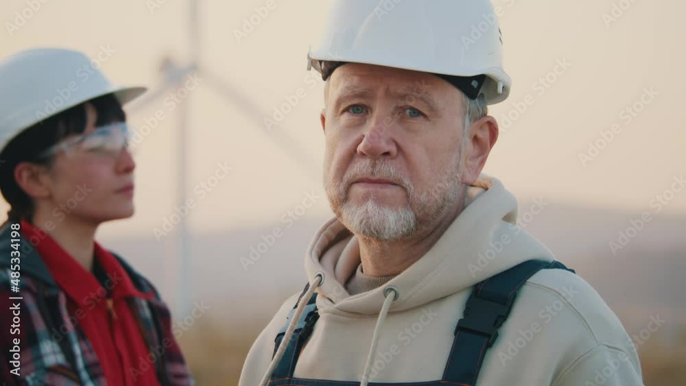 Portrait mature man in hard hat stand look at camera serious outdoor on ecological station. On background woman engineer. Wind turbines rotating at sunset. Slow motion Portrait mature man in hard hat stand look at camera serious outdoor on ecological station. On background woman engineer. Wind turbines rotating at sunset. Slow motion